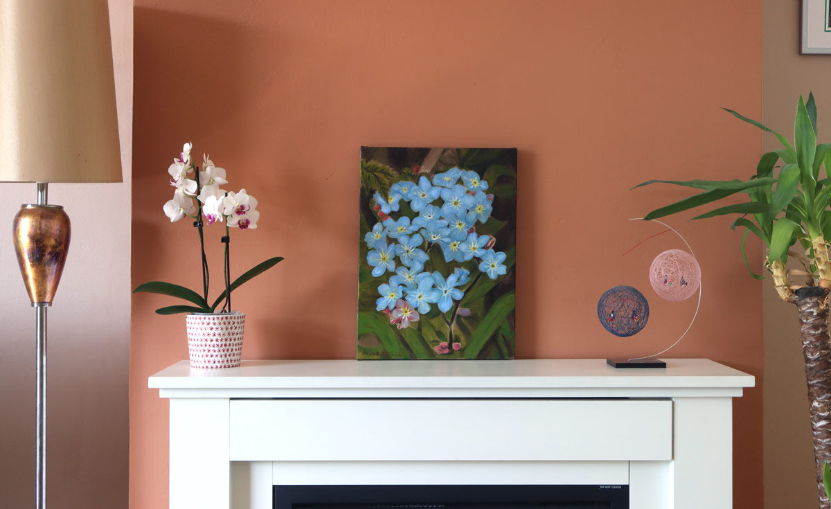A vertical canvas painting of blue forget-me-nots in a vase, displayed on a white fireplace mantel in a styled room with a terracotta wall, a lamp, a potted orchid, and a plant.
